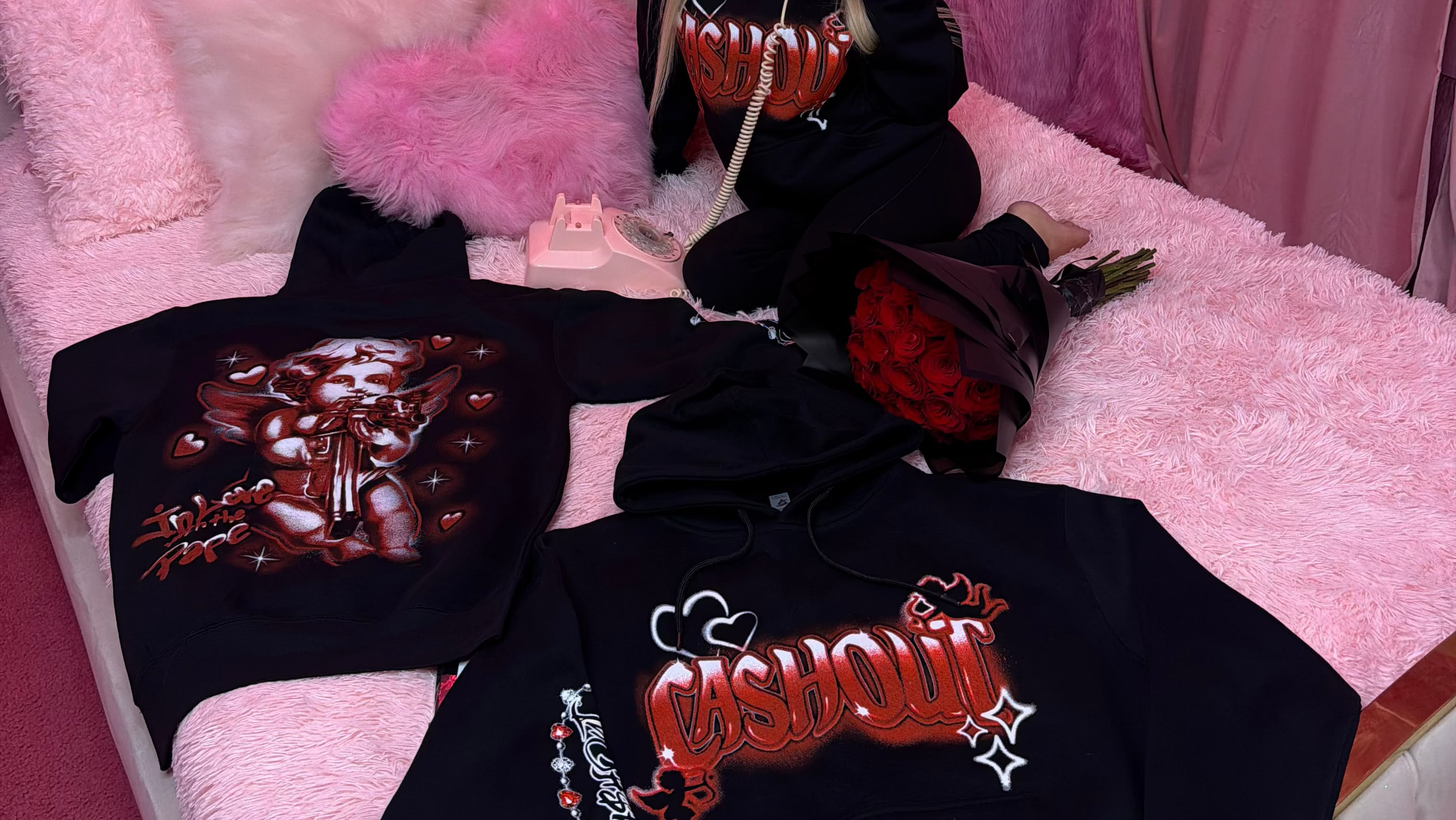 Person sitting on a pink bed with black hoodies featuring red text, surrounded by roses.